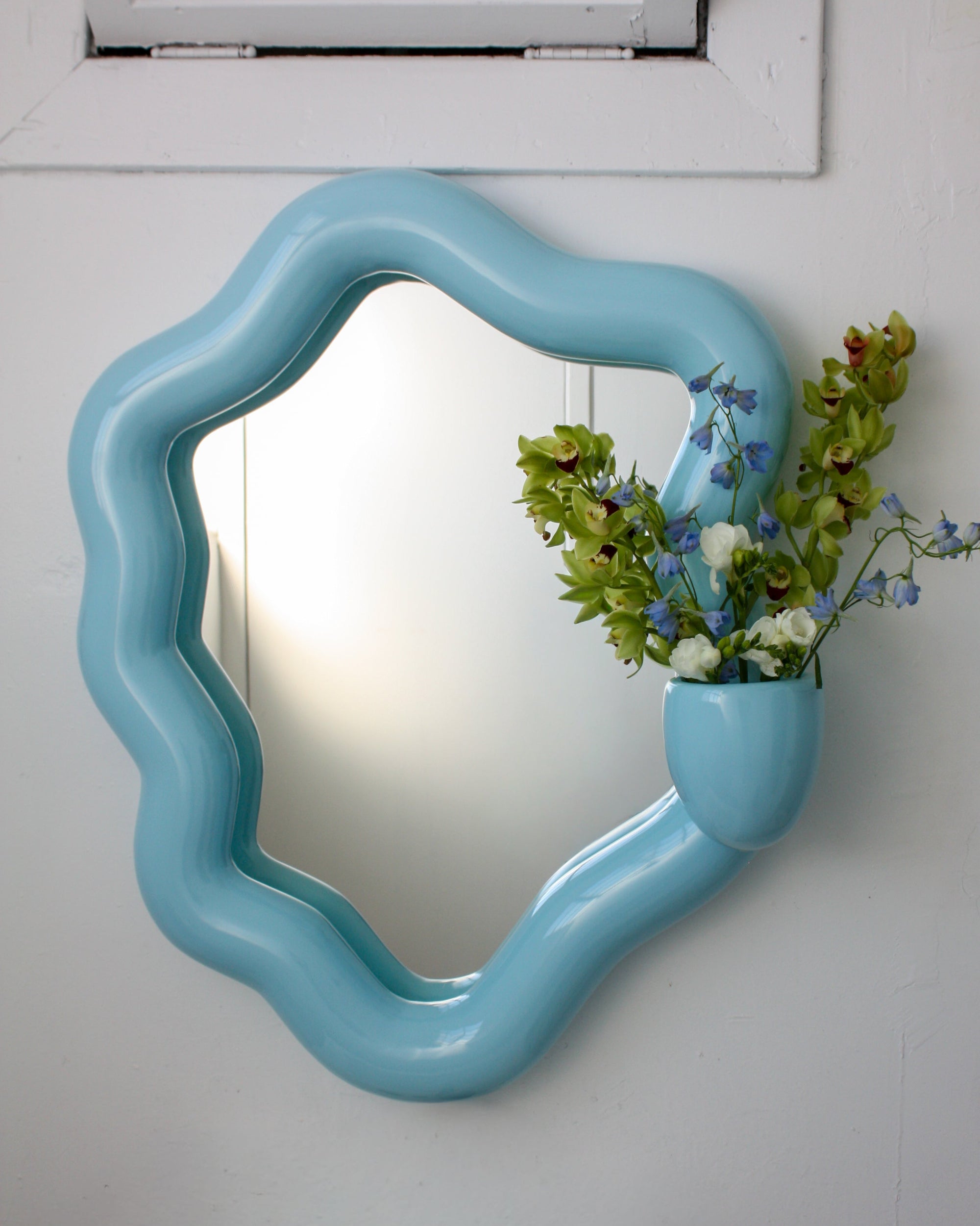 Blue wavy mirror with a vase of flowers on a white wall